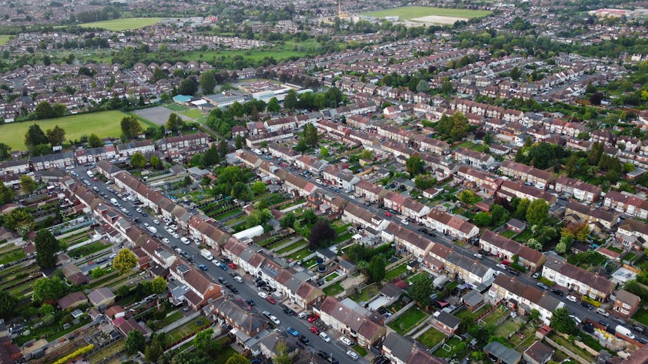 An aerial view of a residential suburban area showing rows of terraced houses with pitched roofs, neatly maintained front gardens, and narrow streets lined with parked cars. The front gardens feature